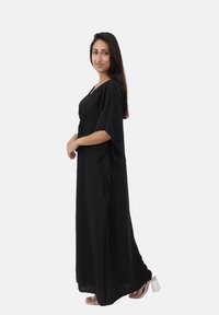 Robe maxi noire avec manches larges, décolleté en V, taille cintrée et jupe fluide. Tissu doux et design longueur cheville. Sandales à talons transparents.