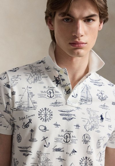 Νέος άνδρας με ανοιχτό καστανά μαλλιά που φοράει ένα λευκό μπλουζάκι πόλο με ναυτικά σχέδια και κείμενο σε ναυτικό μπλε χρώμα.