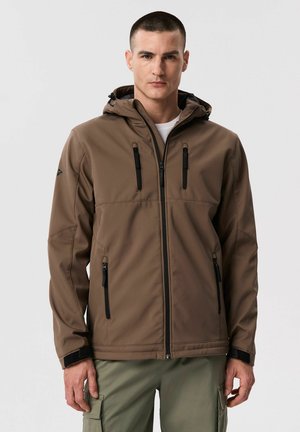 Mężczyzna w brązowej kurtce na zamek błyskawiczny z kapturem i czarnymi kieszeniami na zamek, w zielonych spodniach typu cargo, stojący na jednolitym tle.