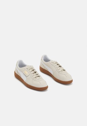 Paire de baskets Puma beige avec une bande latérale blanche et des semelles en caoutchouc marron, vues d’un léger angle en plongée.