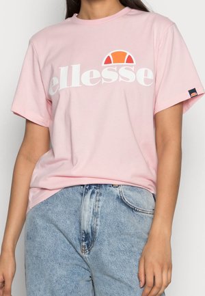 Vrouw draagt een lichtroze t-shirt met wit en oranje "ellesse" logo, gecombineerd met een lichtblauwe, hooggetailleerde spijkerbroek.