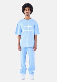 Lichtblauw oversized t-shirt met wit logo en bijpassende blauwe joggingbroek. De stof lijkt zacht; het model draagt witte sneakers.