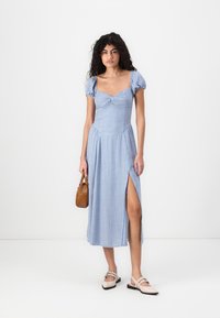 Blå rutete kjole med korte puffermer, tettsittende liv og sideslitt. Kombinert med lyse sko og en liten brun håndveske.