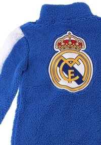 Giacca in pile blu con colletto alto, caratterizzata da maniche bianche e un grande logo ricamato del Real Madrid con corona sulla schiena.