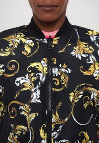 Black bomber jacket with yellow and white baroque floral print, zipper closure, and black ribbed collar. Features logo detail in golden text.
