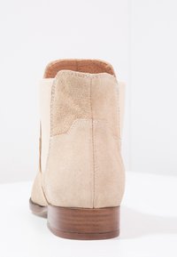Botín de tobillo de ante beige con paneles laterales elásticos y un tacón de madera mediano. Presenta una textura suave y un diseño minimalista.