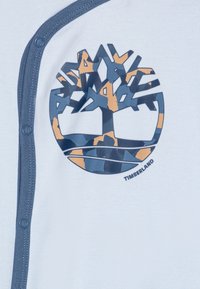 Camisa de algodón blanca con un ribete azul y cierre a presión. Presenta un gran logo de árbol en camuflaje azul y naranja. El texto dice "TIMBERLAND."
