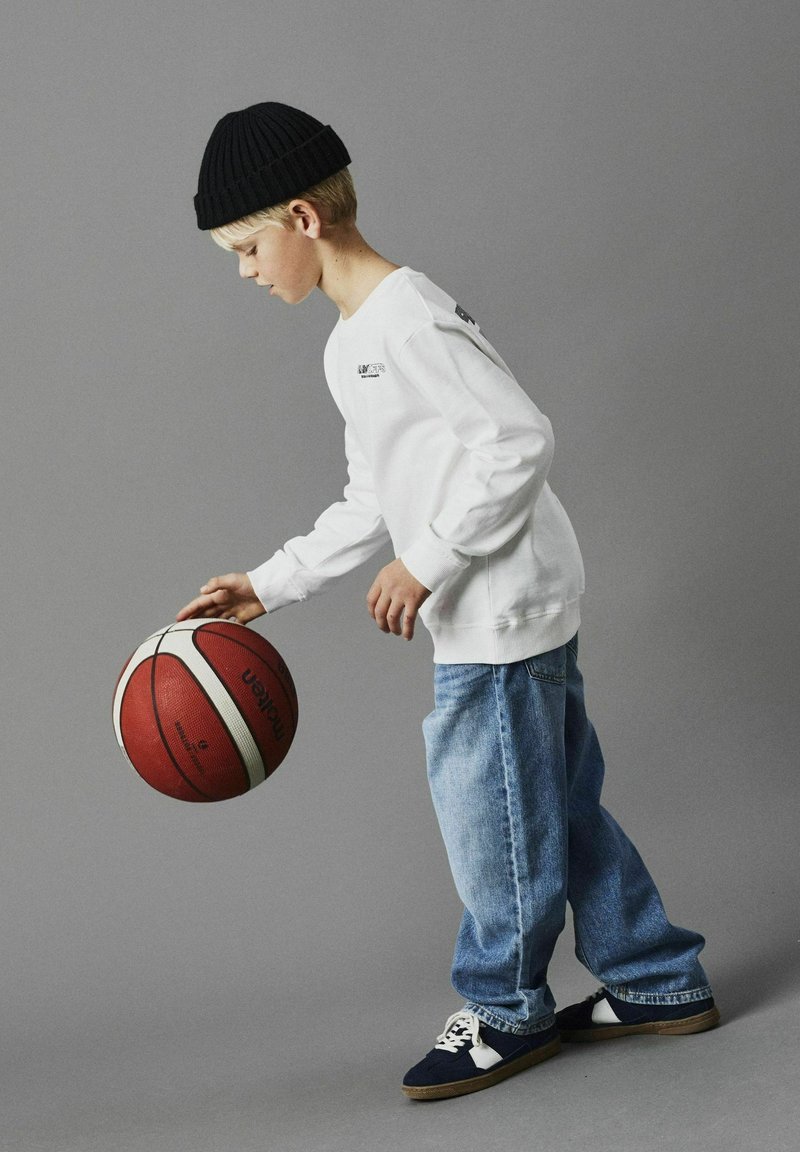 Witte lange mouwen shirt, blauwe spijkerbroek, zwarte beanie en blauwe sneakers. Een rode basketbal wordt in één hand vastgehouden. Grijze achtergrond.