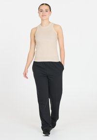 Beige, geripptes Tanktop mit rundem Ausschnitt, kombiniert mit lockeren schwarzen Hosen. Modell trägt schwarze Sneakers, Hände in den Taschen, neutraler Hintergrund.