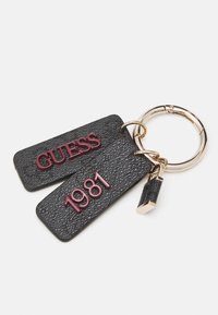 Porte-clés avec deux étiquettes noires texturées, l’une embossée avec "GUESS" et l’autre avec "1981,", attachées à un anneau métallique de couleur dorée.