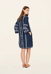 Rochie bleumarin cu broderie albă pe mâneci și pe spate, având o croială lejeră și buzunare; textură de țesătură netedă.