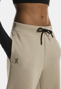 Beige sweatpants med en snøre i taljen med en sort detalje, sidelommer og et logo trykt på venstre lår. Glat stoftekstur.