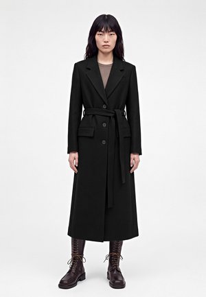 Femme aux longs cheveux noirs portant un long manteau noir ceinturé, des bottines marron lacées et un haut marron, debout devant un fond uni.