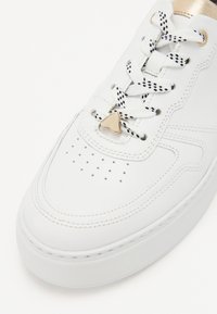 Valge nahast sneaker, millel on augustatud varvas, tekstuursed detailid, mustvalged mustriga paelad ja väike kuldne kolmnurkne detail paelte peal.