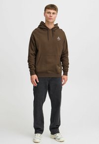 Brauner Kapuzenpullover mit Kordelzug, Bauchtasche und Logo-Detail; kombiniert mit dunklen Jeans und weißen Sneakern. Das Modell steht frontal.