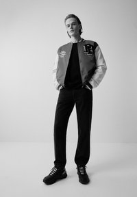 Veste universitaire rouge et blanche, t-shirt noir, jean noir et baskets noires avec lacets. La veste est ornée d'un logo et de détails brodés.