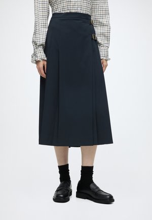 Femme portant une jupe midi bleu foncé avec des boucles latérales, des chaussettes noires, des mocassins noirs et un chemisier à manches longues à carreaux avec des poignets volantés.