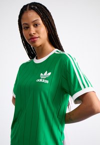 Camiseta deportiva verde con rayas verticales, cuello y acentos en las mangas blancos, cuenta con el logo de Adidas en la parte delantera. Fabricada con tela transpirable.