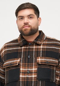Brown and black plaid flannel shirt with four front pockets, button-up closure, and a collared neck. Soft texture and warm design.