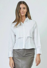 Camicia bianca con un design cropped, caratterizzata da maniche lunghe e un frontale a strati, abbinata a una gonna aderente in pelle grigia.