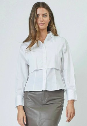White button-up shirt with a cropped design, featuring long sleeves and a layered front, paired with a fitted gray leather skirt.