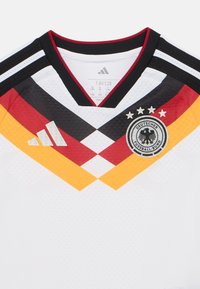 Camiseta nacional de fútbol de Alemania con base blanca y acentos diagonales en negro, rojo y amarillo. Cuenta con un cuello negro y un emblema bordado.
