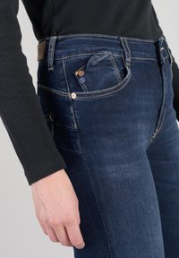 Jeans de un azul oscuro con un diseño ajustado, que cuentan con un bolsillo trasero prominente, costuras en contraste y un pequeño detalle de logo.