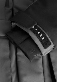 Čierna športová bunda s hladkým, elastickým materiálom. Rukávy majú nastaviteľný suchý zips, s bielou nášivkou "BAKER" na vonkajšej strane rukávu.