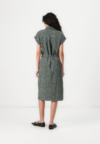 Femme aux longs cheveux bouclés portant une robe midi verte à manches courtes et motifs, ainsi que des chaussures plates noires, debout de dos sur un fond blanc.