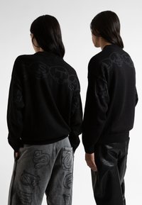 Μαύρο πουλόβερ με τονικό σχέδιο κρανίου στο πίσω μέρος. Συνδυασμένο με γκρι παντελόνι που παρουσιάζει παρόμοιες γραφικές λεπτομέρειες, δημιουργώντας μια ομοιόμορφη εμφάνιση.