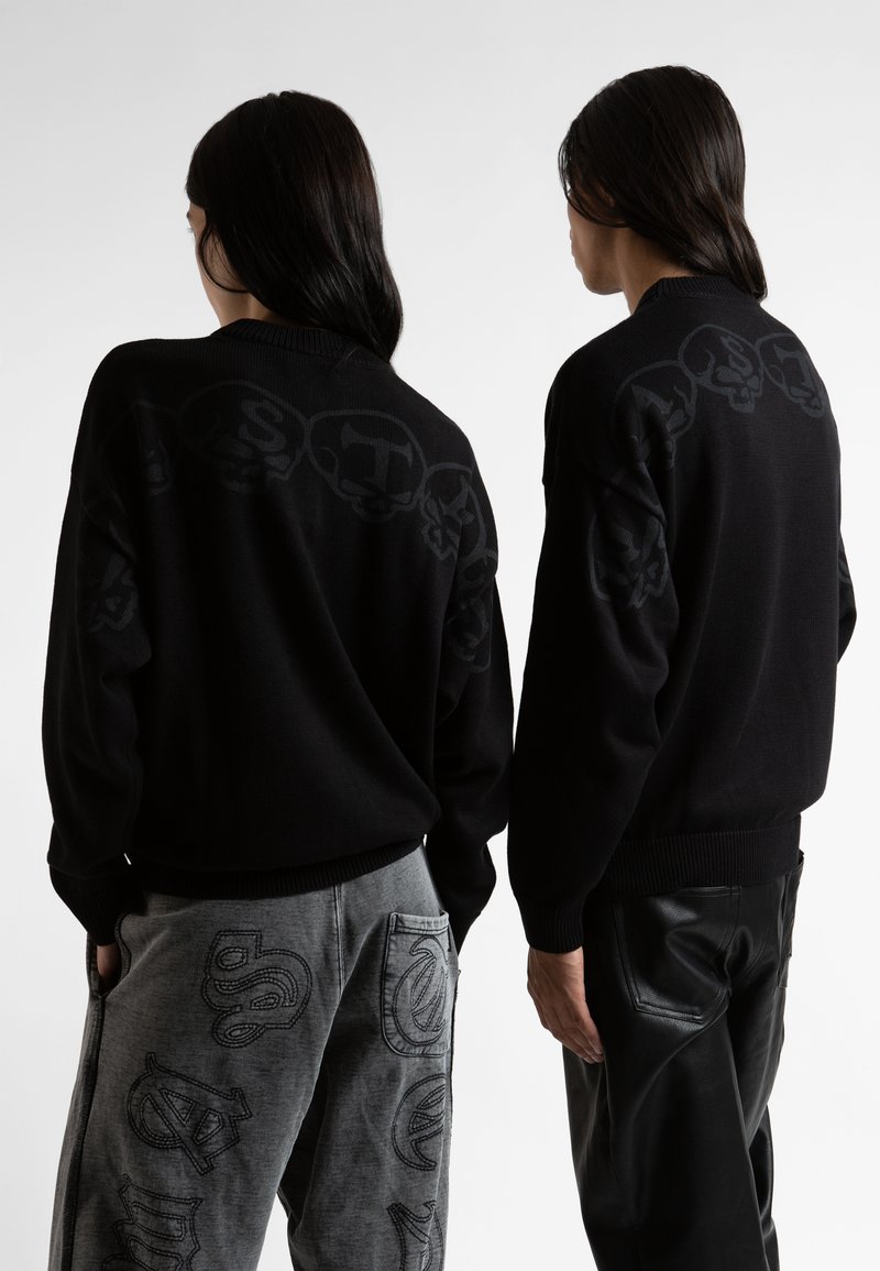 Μαύρο πουλόβερ με τονικό σχέδιο κρανίου στο πίσω μέρος. Συνδυασμένο με γκρι παντελόνι που παρουσιάζει παρόμοιες γραφικές λεπτομέρειες, δημιουργώντας μια ομοιόμορφη εμφάνιση.