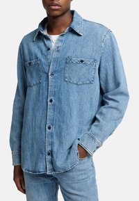 Camicia in denim azzurro chiaro con bottone frontale, due tasche sul petto e dettagli cuciti. Maniche lunghe e design con colletto classico.