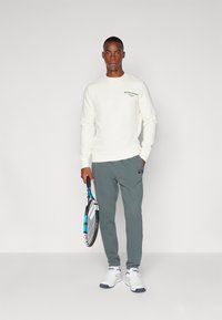 Felpa bianca con logo verde, pantaloni grigi e sneakers bianche. Tiene una racchetta da tennis con dettagli neri e blu. Design semplice.