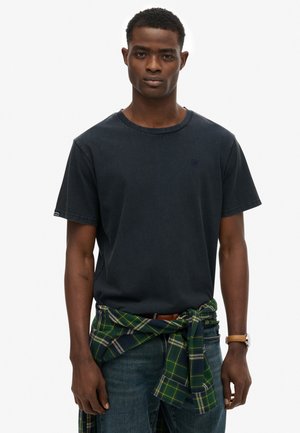 Navyblauwe katoenen t-shirt, ronde hals, korte mouwen, gecombineerd met donkere spijkerbroek en een groen geruit flanellen overhemd om de taille geknoopt.