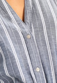 Primer plano de una persona que lleva una camisa de botones de rayas verticales azul y blanca con dos botones visibles abrochados.