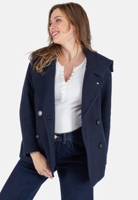 Navyblauw dubbelzijdig jasje met grote knopen, licht getextureerde stof en brede revers, gedragen over een witte top en donkere jeans.