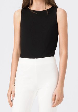 Femme portant un haut noir sans manches associé à un pantalon blanc taille haute sur un fond clair uni.