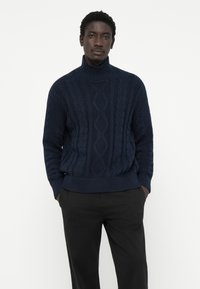 Pulover navy cu guler înalt din tricot cu model texturat și manșete cu nervuri, purtat cu pantaloni negri.