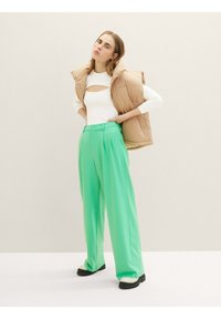 Beige gepolsterte Weste, weißes langärmliges Oberteil mit Aussschnitt und grüne weite Hosen mit Falten, kombiniert mit schwarzen und cremefarbenen Plateauschuhen.