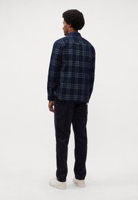 Man met afro kapsel, gekleed in een marineblauwe geruite blouse, donkere corduroy broek en witte sneakers, staande met zijn rug naar de camera op een witte achtergrond.