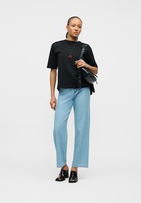 Γυναίκα που στέκεται φορώντας μαύρο τοπ, ανοιχτό μπλε φαρδιά τζιν, μαύρα παπούτσια με τακούνι, κολιέ με μενταγιόν τριαντάφυλλο και κρατώντας μαύρη τσάντα ώμου.