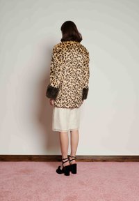 Mulher de costas para a câmara, a usar um casaco de pêlo com padrão de leopardo com punhos de pêlo escuro, saia branca e sandálias pretas de salto alto sobre um tapete cor-de-rosa.