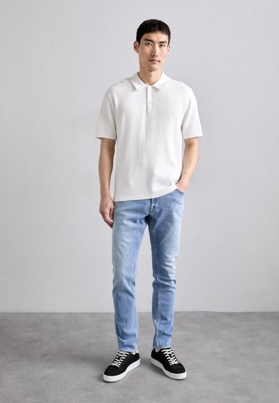 Witte polo met korte mouwen en een gestructureerd patroon, gecombineerd met lichtblauwe jeans en zwarte sneakers met witte zolen.