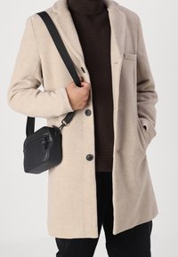 Beige wool coat with a tailored design, black buttons, and a single chest pocket, worn over a dark brown ribbed sweater, paired with a small black bag.