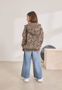 Geacă cu glugă cu imprimeu de leopard, în culori bej și maro, asortată cu pantaloni denim largi de culoare albastru deschis. Pantofii sport albi completează ținuta.