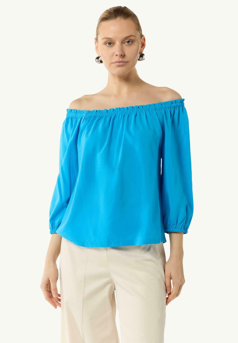 Jasnoniebieska bluzka typu off-the-shoulder o luźnym kroju, z długimi bufkami i elastycznym dekoltem. Gładki materiał w połączeniu z kremowymi spodniami.