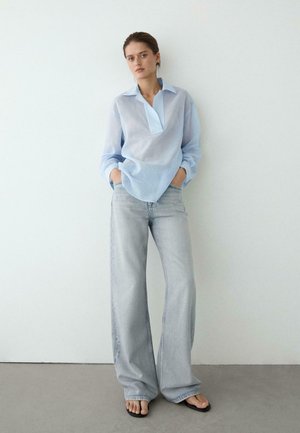 Eine Frau steht an einer weißen Wand und trägt eine lockere, hellblaue Bluse, weit geschnittene, ausgewaschene Jeans, die Hände in den Taschen und schwarze Sandalen.