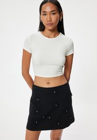 Weißes kurzärmeliges Crop-Top, kombiniert mit einem schwarzen Minirock, der mit mehreren kleinen schwarzen Schleifen-Applikationen geschmückt ist, die mit Perlen verziert sind. Gehobene Texturen.