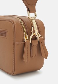 Hnědá texturovaná kožená taška v rohu s dvojitými zlatými zipy a zlatým zámkem s vyrytým nápisem "Valentino" na bílém pozadí.