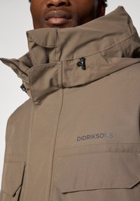 Brun vandtæt jakke med høj krave, der har lynlåse og to forlommer. Brandlogoet "DIDRIKSONS" vist på brystet.